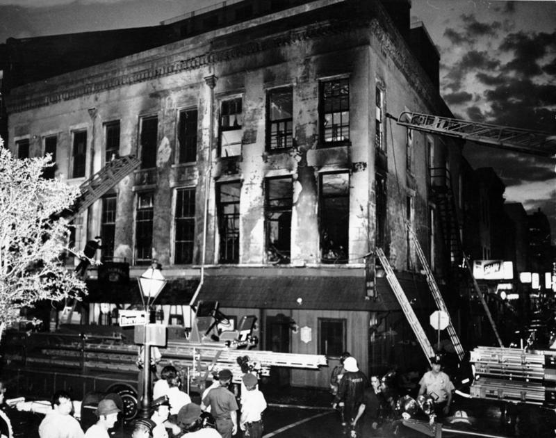 A black and white photo shows a large, damaged building at night. Fire trucks are parked outside, with ladders extended to the upper floors. Firefighters and police officers are gathered around the scene, working under the streetlights.
