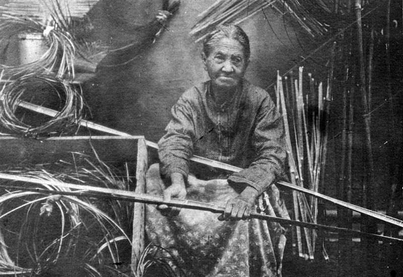 An elderly woman sits among bamboo tools and materials, holding a long piece of bamboo. She wears a patterned skirt and a long-sleeved top, with her hair tied back. The background contains various bamboo sticks and woven items.