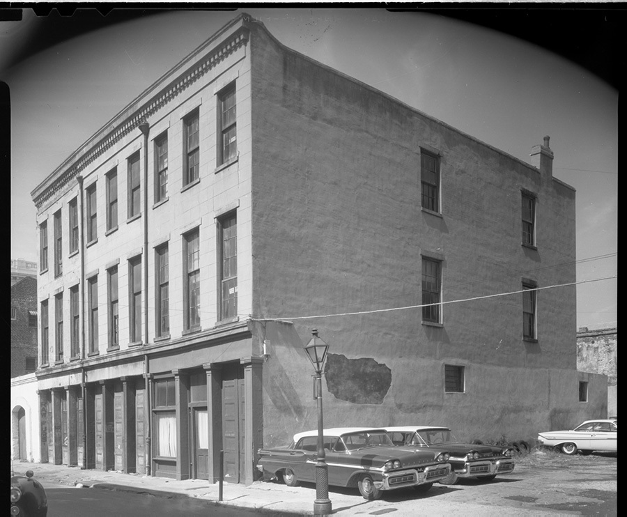 A black and white photo of a three-story building with large windows. Classic cars are parked along the street beside it. The scene has a vintage, mid-20th century urban feel.