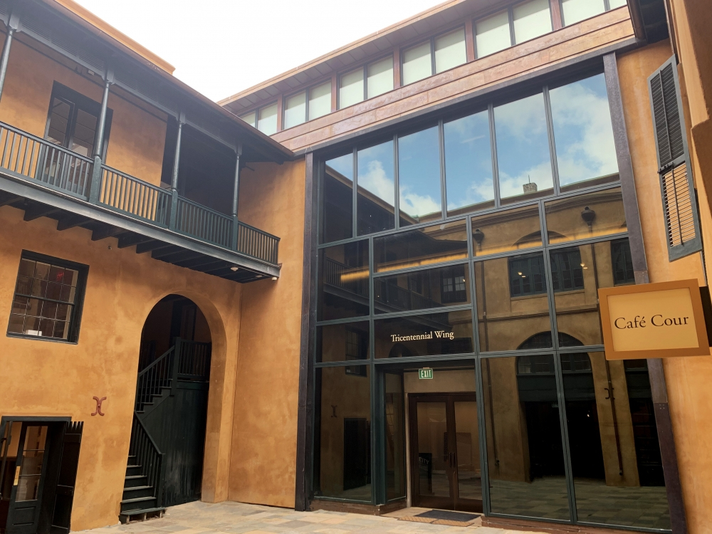 A modern courtyard with light brown walls features a glass entrance labeled Ticonderoga Wing. A sign for Café Cour is visible on the right. Theres a staircase on the left, and reflections of the sky and clouds can be seen in the glass.