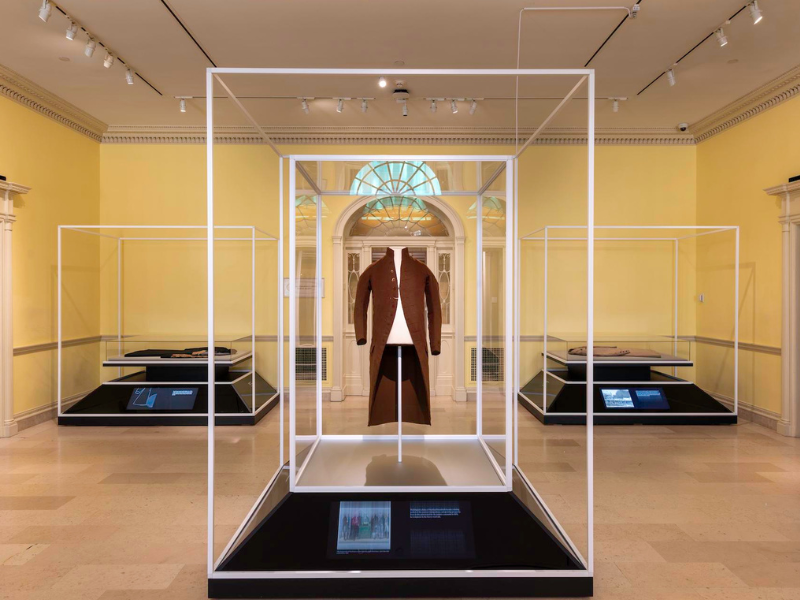 A brown coat displayed in a glass case in a museum gallery. The room has yellow walls, and additional exhibits are visible in the background. The ceiling has spotlights illuminating the displays.
