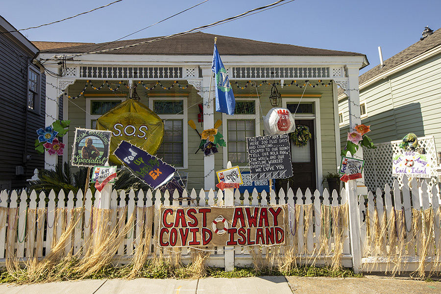 A house decorated with various signs and flags, featuring messages like Cast Away Covid Island and S.O.S. The porch has colorful flowers and lights, with a fence surrounded by string decorations and artwork.