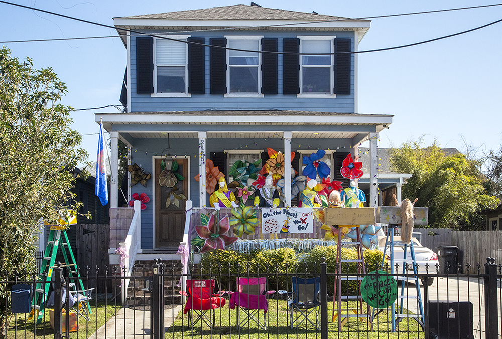 A two-story house decorated with colorful pinwheels, flowers, and banners. A sign reads Who Dat Parade Just Roll!!. A fenced yard contains folding chairs and a green ladder. .