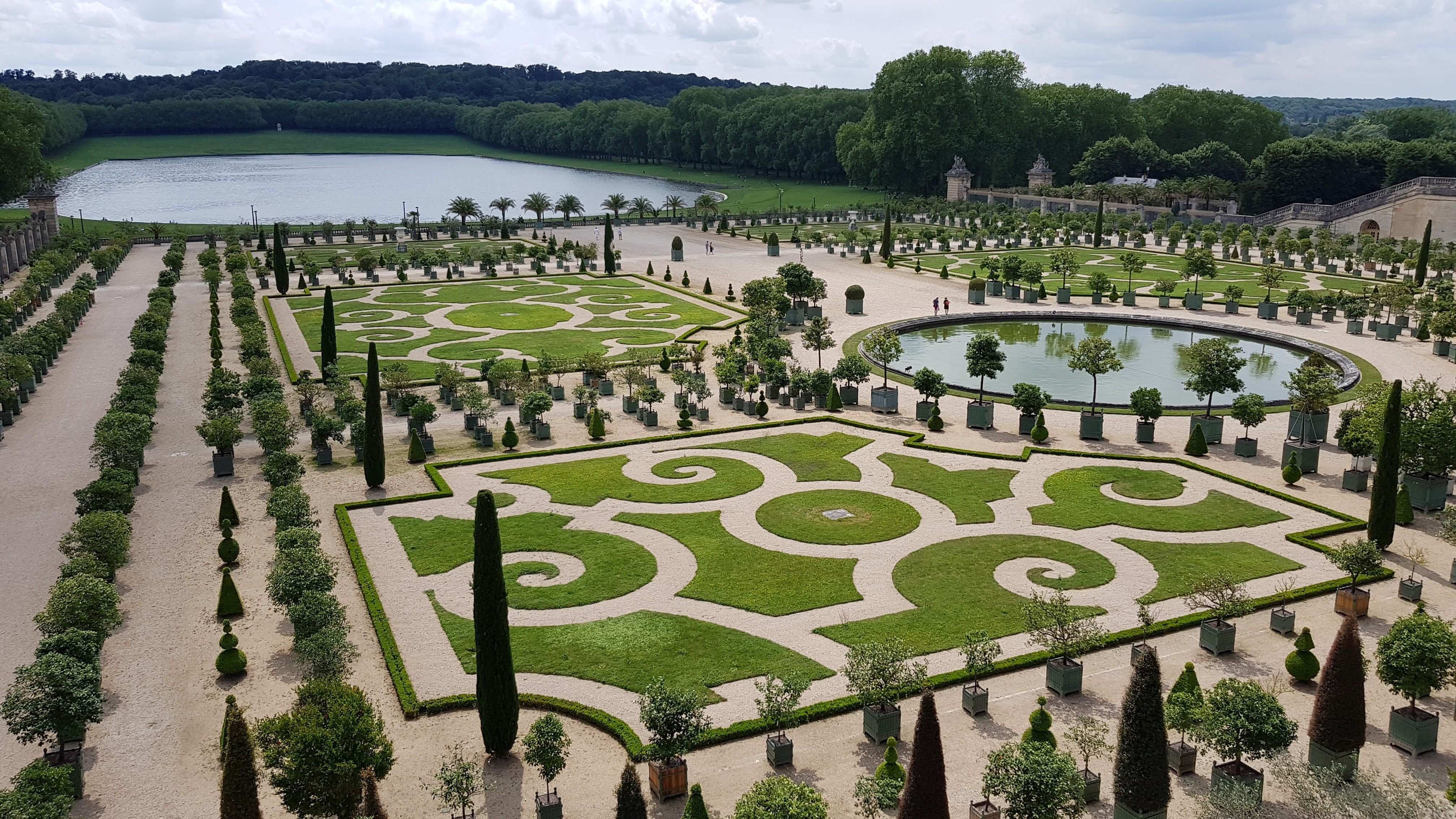 Ornate gardens with symmetrical hedges and pathways, featuring a circular pond. The gardens are surrounded by dense trees, with a large lake in the background under a partly cloudy sky.