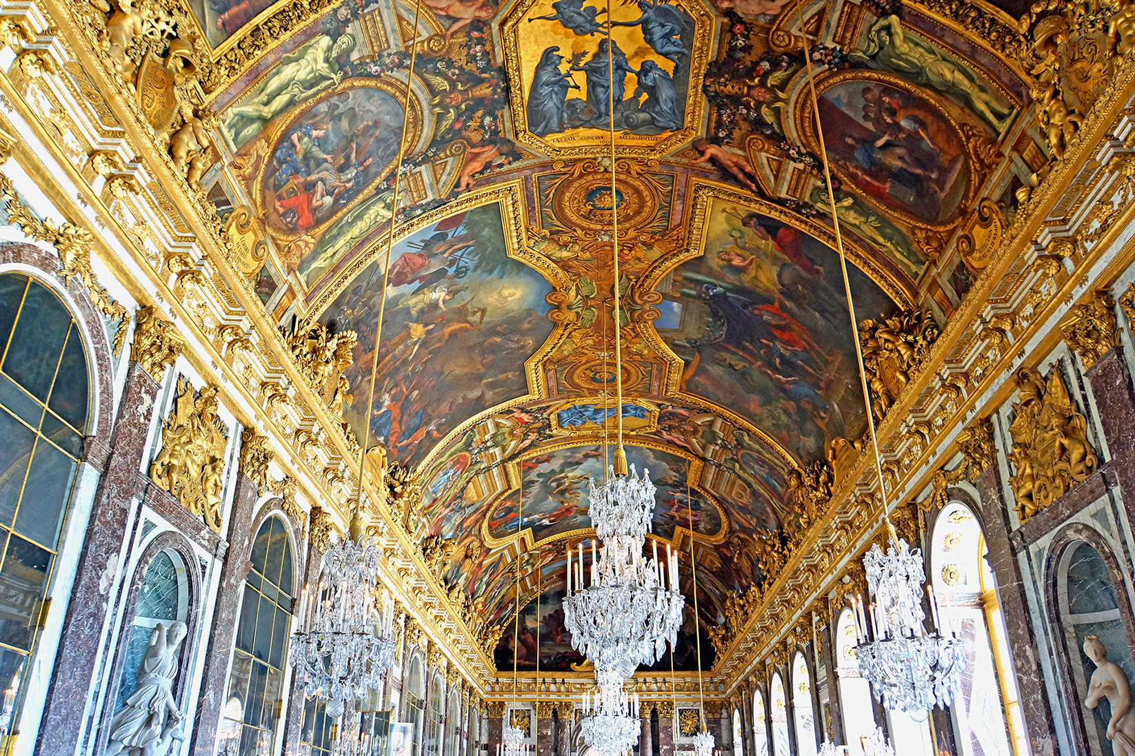 The image shows the Hall of Mirrors in the Palace of Versailles, featuring an ornate ceiling with elaborate paintings and chandeliers hanging from the intricate designs. Tall arched windows line the walls, reflecting light onto the lavish interior.