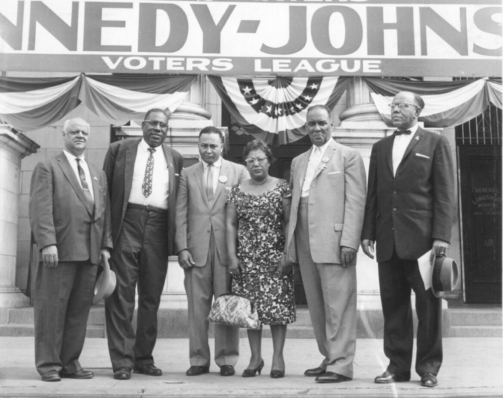 A group of six individuals, five men and one woman, stand on the steps of a building with a Voters League sign above. They are dressed formally, and the building is adorned with patriotic bunting.