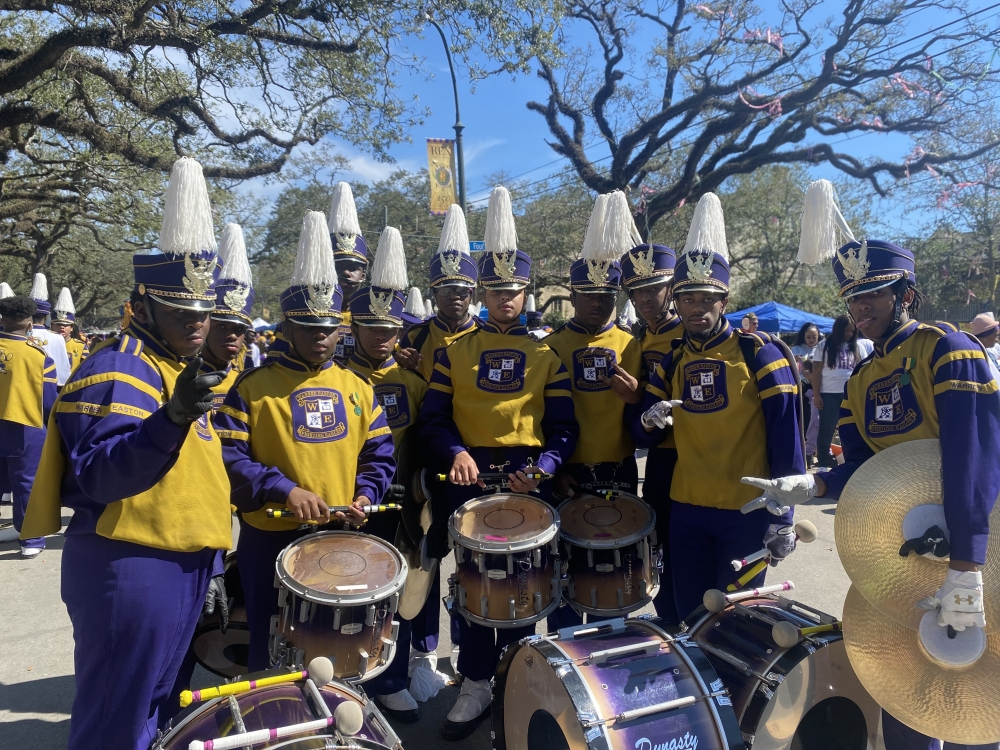 A marching band dressed in purple and yellow uniforms gathers around a set of drums. They wear tall, feathered hats and stand under leafy trees on a clear day.