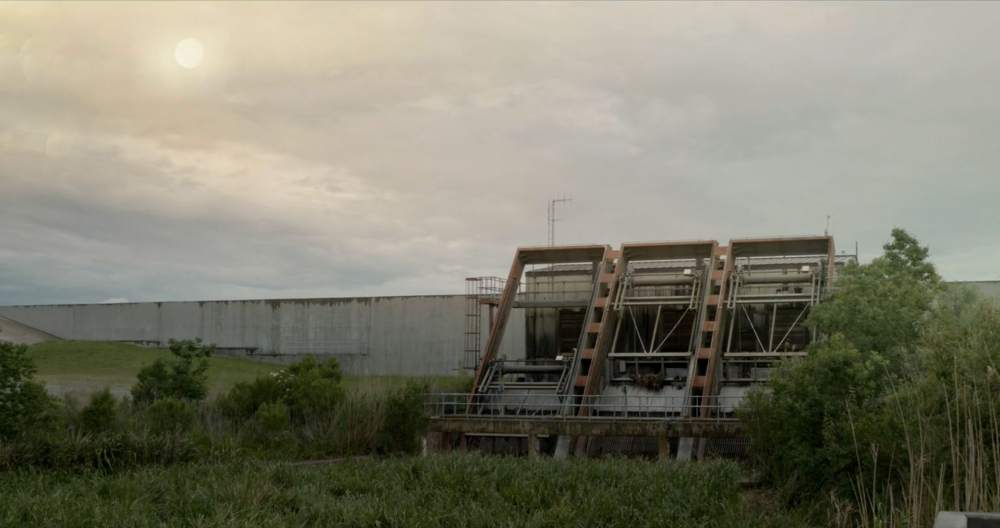 A large industrial structure with machinery and metal framework sits near lush greenery and tall grasses. A massive concrete wall stretches across the background under a cloudy sky with a partially visible sun.
