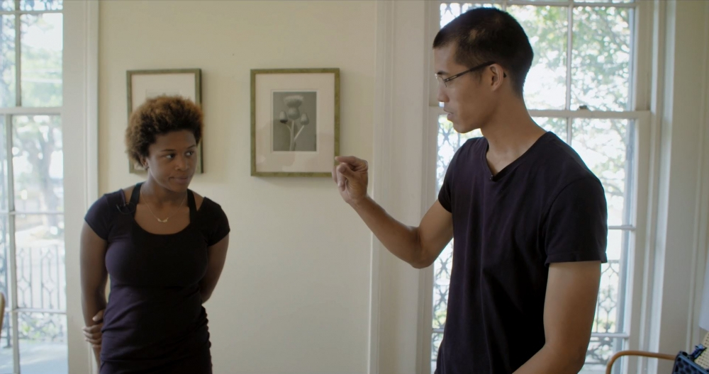A man and woman stand in a room with large windows and framed photos on the wall. The man gestures with his hand as the woman looks on with a thoughtful expression. Theyre both wearing black shirts.