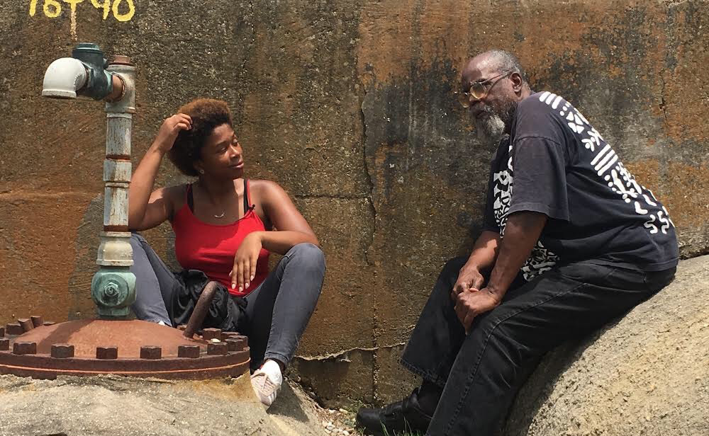 A young woman in a red top and jeans sits on the ground near a metal pipe, talking to an older man in a black shirt and glasses who is seated on a rock. They appear to be having a casual conversation. A concrete wall is in the background.