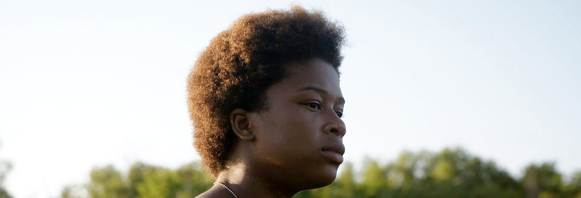 Person with curly hair looking thoughtfully into the distance, set against a clear sky and trees in the background.