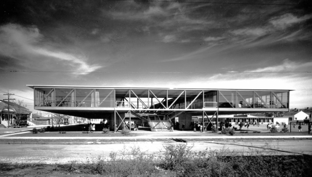 Black and white photo of a modernist building with a flat roof, large glass windows, and an elevated structure on stilts. A central staircase leads up, and people are gathered around the entrance. The sky is partly cloudy.