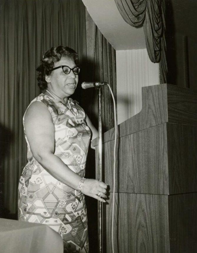 A woman wearing glasses and a patterned dress speaks at a podium with a microphone in front of her. The setting appears to be indoors, with curtains in the background. The image is in black and white.