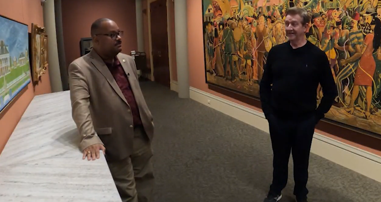 Two men stand in front of a large painting, engaged in conversation.