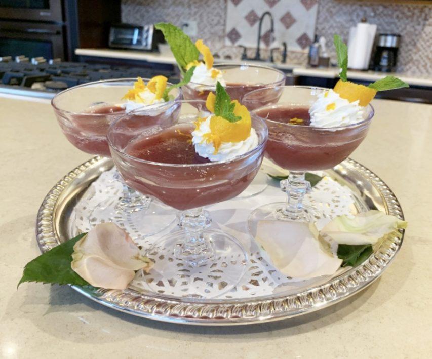 Four dessert glasses filled with a dark red gelatin dessert topped with whipped cream, orange slices, and mint leaves. They are arranged on a silver tray with lace doily and white rose petals.