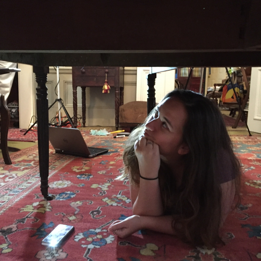 A woman lying on a patterned rug under a table, looking thoughtfully upwards with her hand on her chin. Various objects, including a laptop, are scattered across the floor.