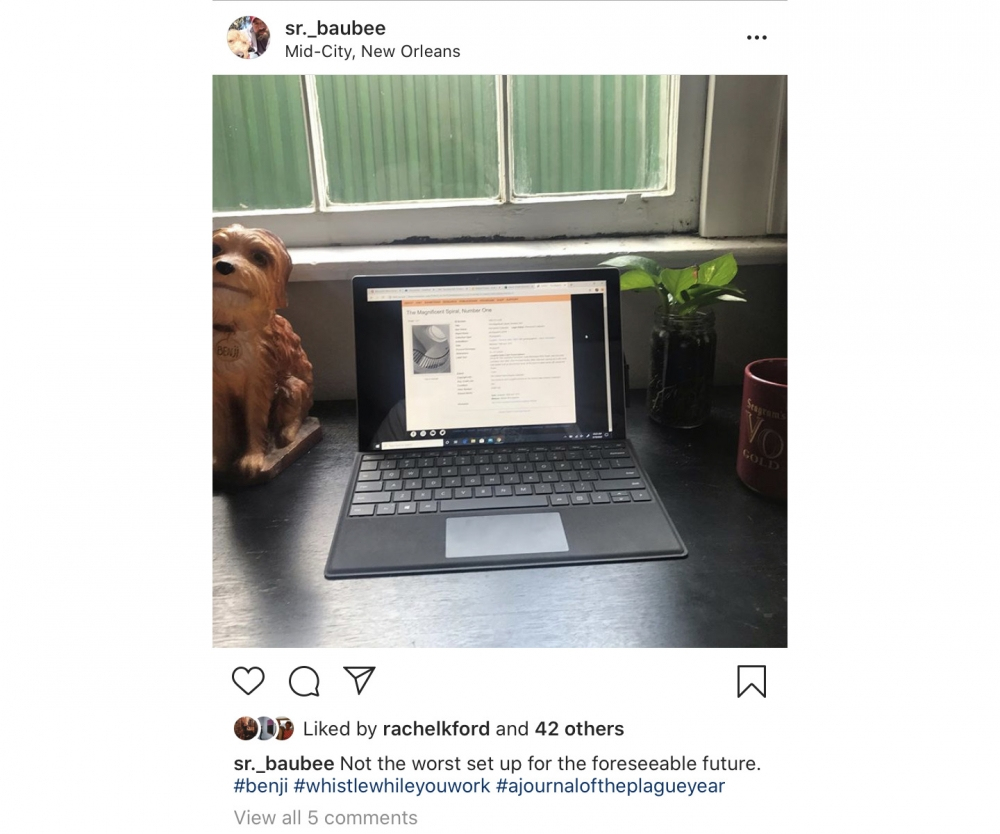 A tablet with a keyboard sits on a table under a window. A small statue of a dog and a potted plant are next to it. The screen displays a document. The caption mentions working remotely and hashtags like #benji and #whistlewhileyouwork.