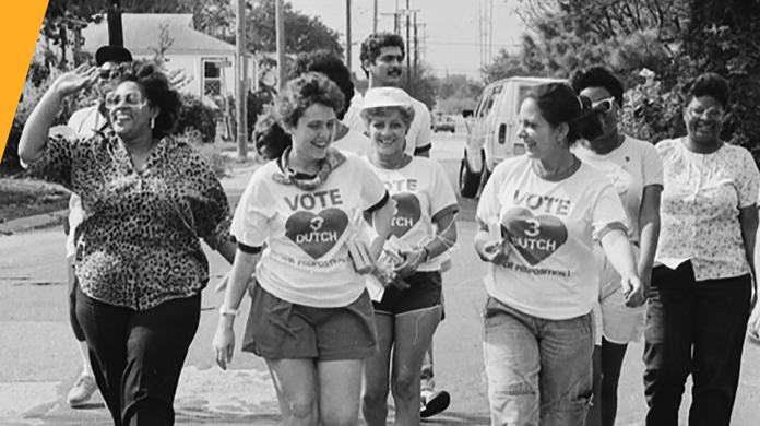 A group of people walk energetically down a street, some wearing Vote 3 Dutch shirts. The mood is lively as they engage in what appears to be a campaign event or community march, set against a backdrop of residential houses and trees.