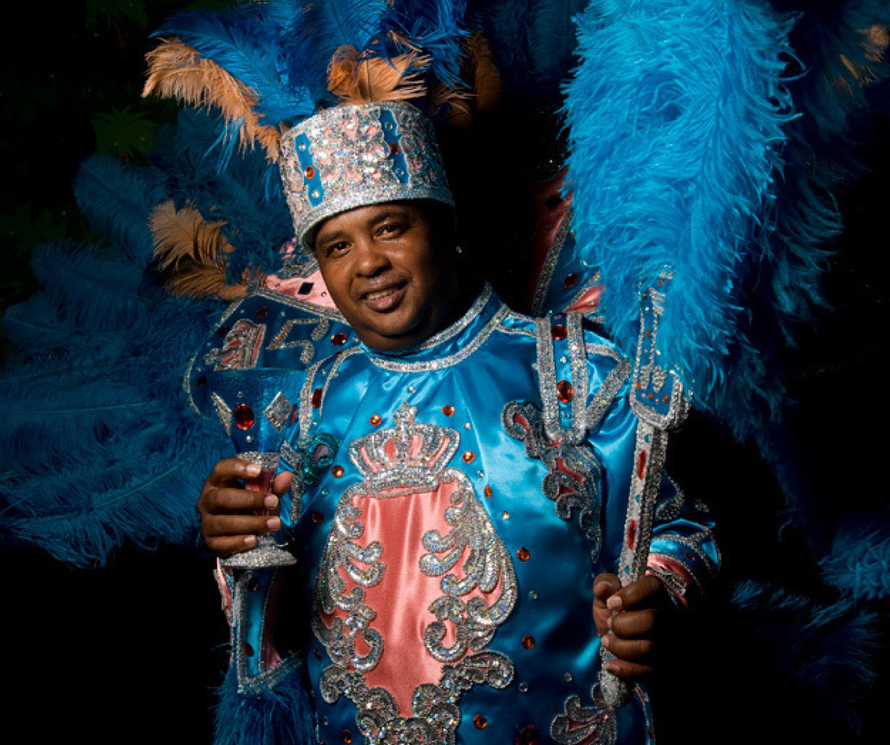 A person dressed in an elaborate blue and pink costume adorned with sequins and feathers, holding a bejeweled staff and a drink. The headdress features large blue plumes, and the outfit sparkles under the light.