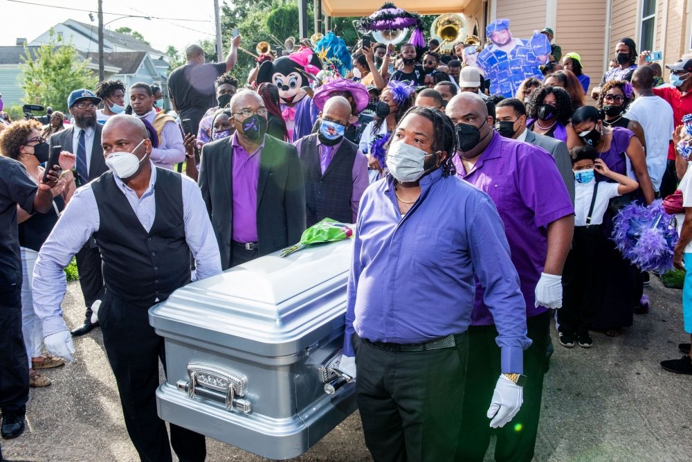 A group of men in purple shirts and black vests carry a silver casket. They are followed by a brass band and people dressed in purple, celebrating with balloons and costumes. All participants wear face masks. The scene is lively and colorful.