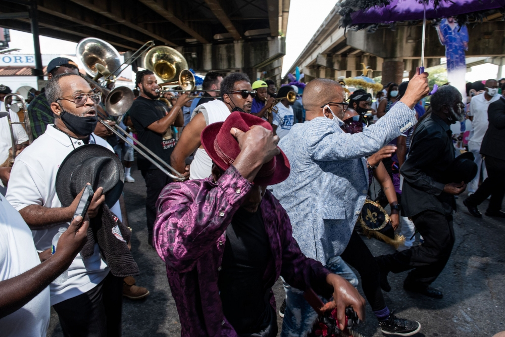 A lively outdoor scene with a brass band and people dancing under a bridge. Musicians play trombones while participants in colorful outfits and hats celebrate together. The atmosphere is festive and energetic.