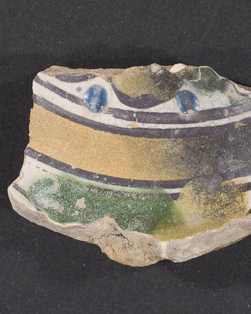 A fragment of ancient pottery featuring a colorful pattern with blue, yellow, and green curved lines on a black background. The edges are worn and uneven, revealing the texture of the ceramic material.