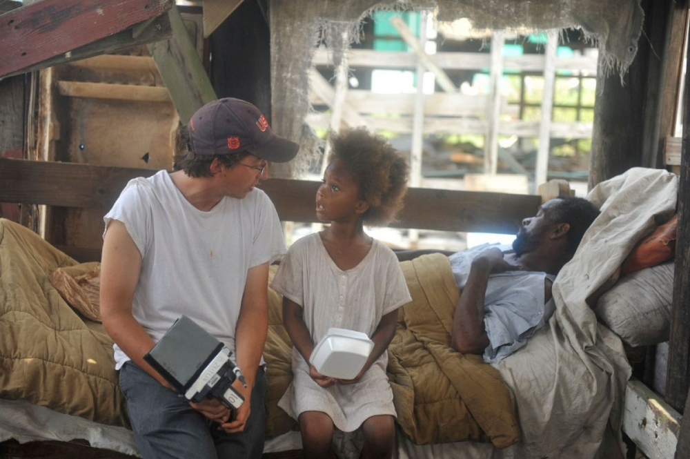 A person in a cap sits next to a child holding a container, talking to a man lying on a bed in a rustic setting. The background features wooden structures and a burlap covering.