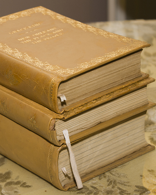 Three stacked vintage books with ornate gold details on the spines, resting on a patterned fabric surface. The books have aged, beige covers and visible ribbons marking pages.