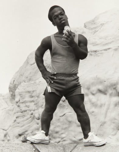 A man in athletic wear stands on a rock with one hand on his hip and the other holding an ice cream cone. He is wearing a tank top, shorts, and sneakers. The background features large rock formations.