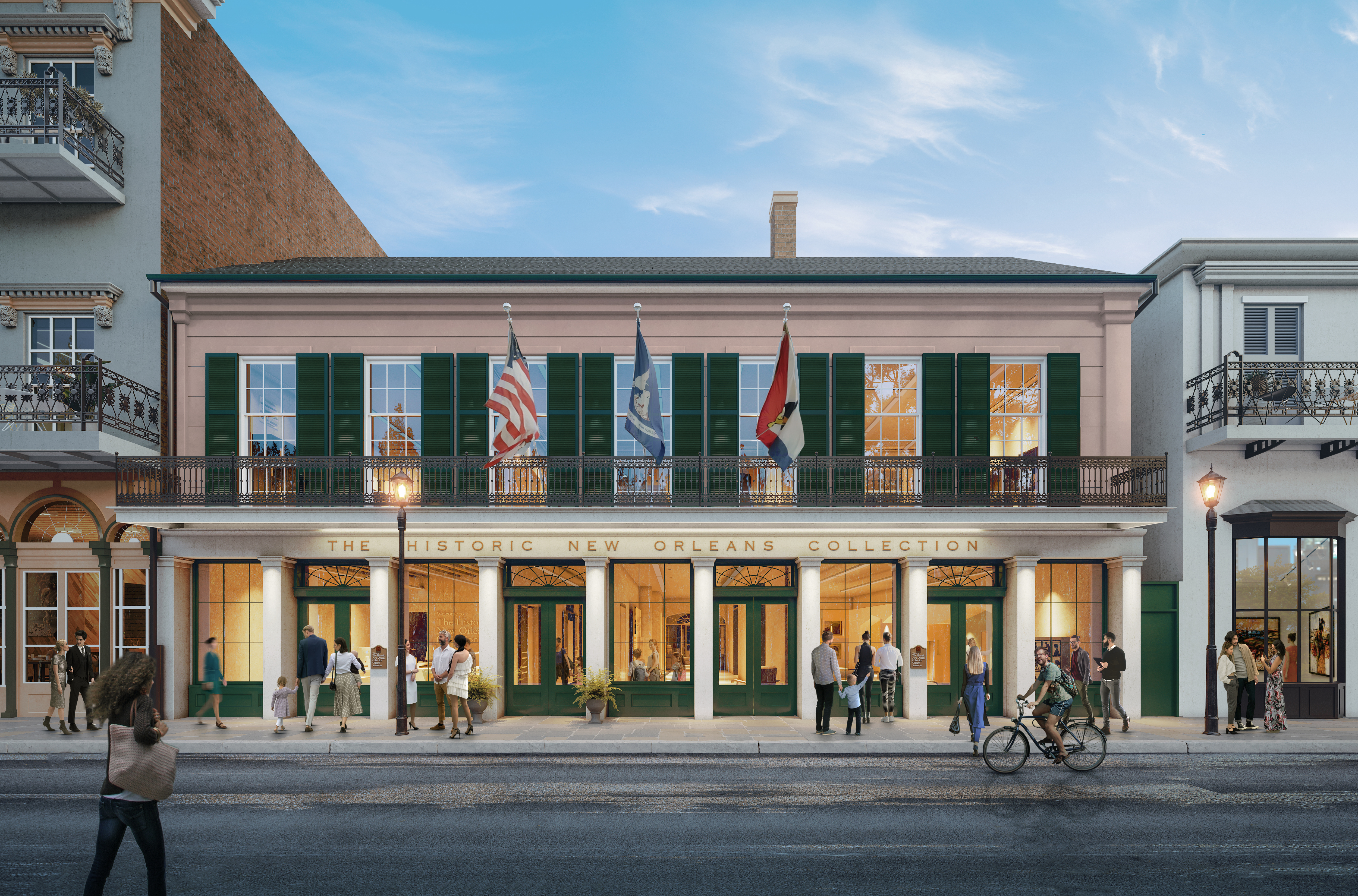 A historic building in New Orleans features a pink facade with green shutters. Three flags fly above the entrance. People walk and stand in groups outside, and a person rides a bicycle past on the street. The sky is clear and blue.