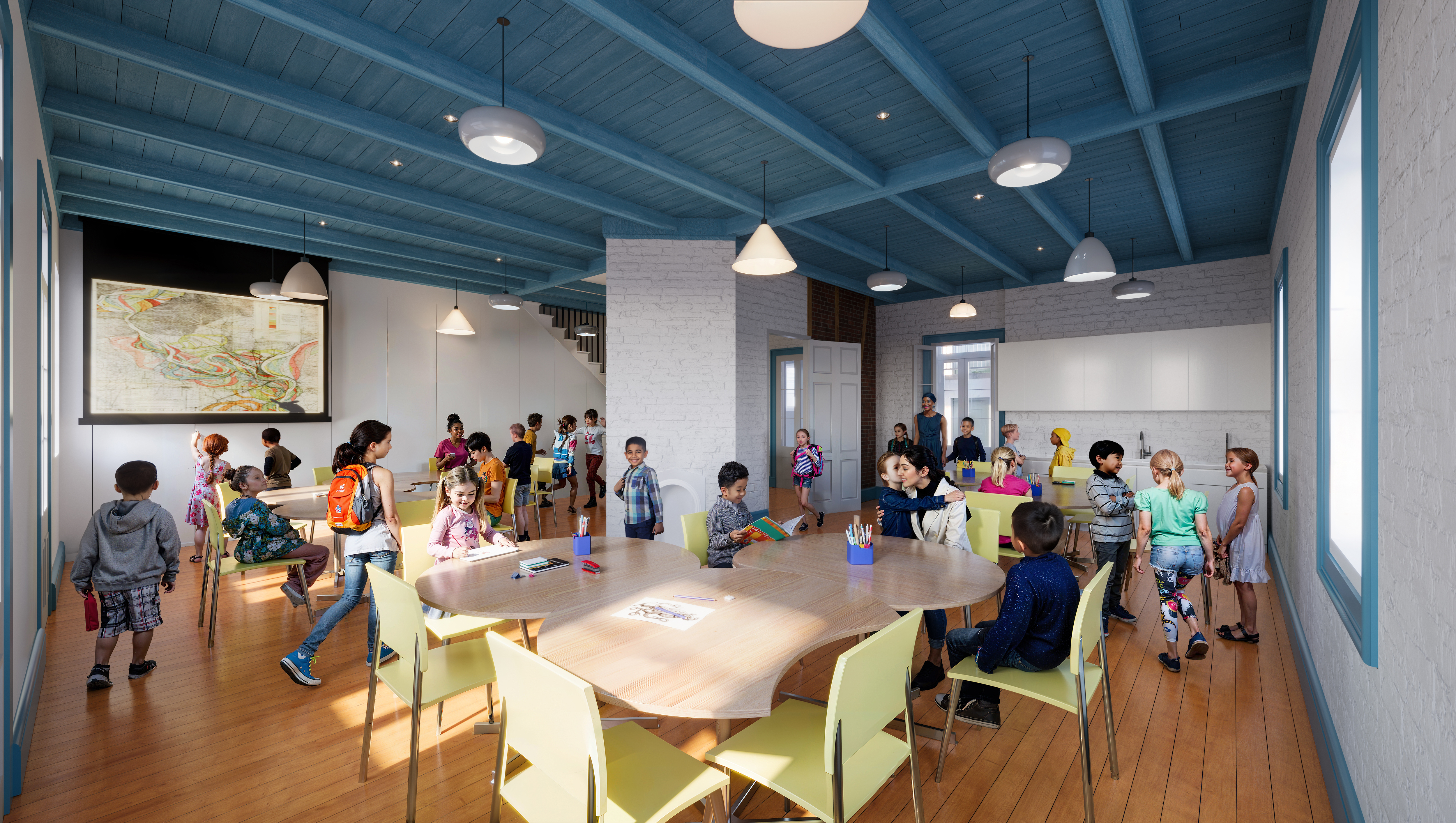 A bright classroom with a group of children gathered around tables. The room has blue ceiling beams, wooden floors, and white walls. A large map hangs on the wall, and pendant lights illuminate the space. Children are engaged in various activities.