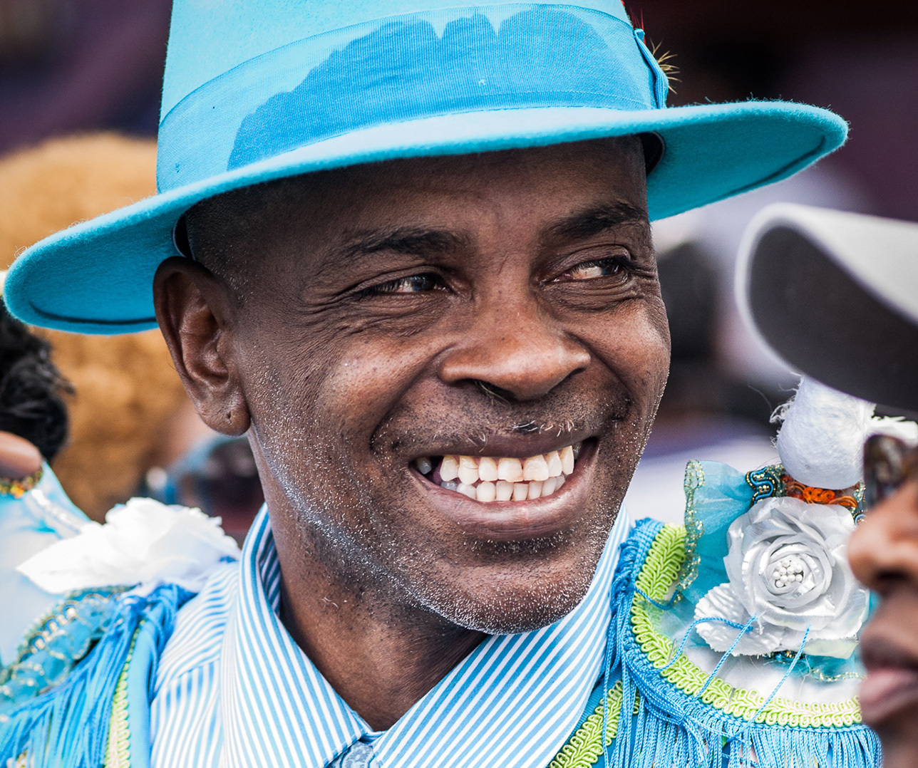 A person wearing a bright blue hat, embellished with a white flower and a feather, smiles widely. Their outfit features intricate beadwork and stripes. The background is blurred, with another person partially visible.