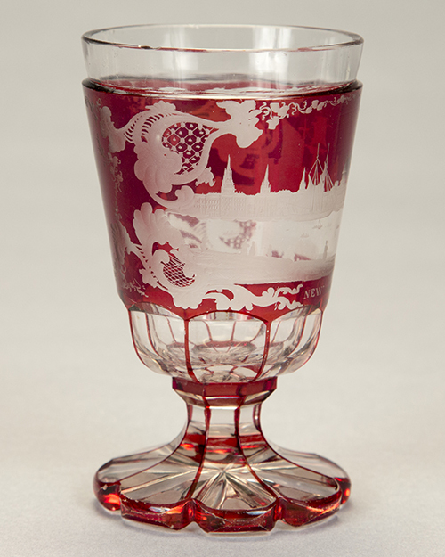 A decorative, vintage glass goblet with a red tint and intricate designs, including buildings, engraved on its surface. The goblet features a textured, flared stem and a scalloped base.