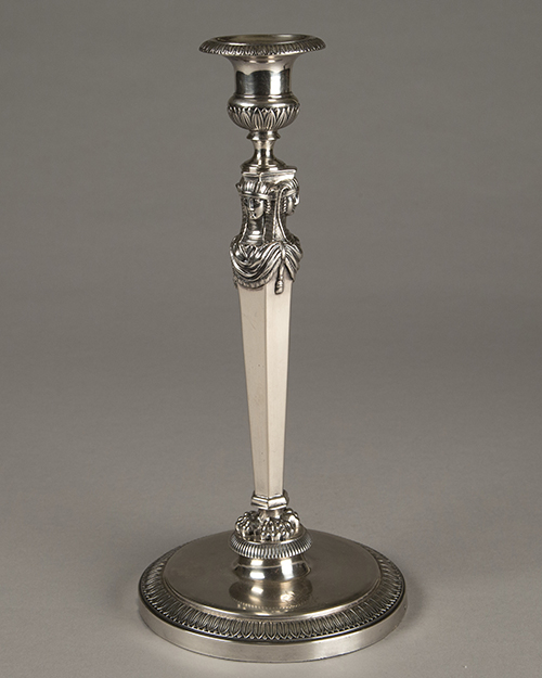 A silver candlestick with an ornate, decorative column and a round base, set against a plain gray background. The intricate design features carved elements near the top and bottom.