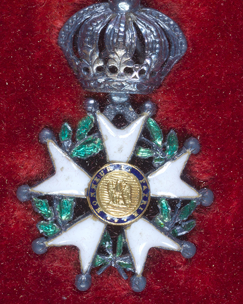 A medal with a gold central emblem surrounded by a blue ring with silver lettering. The medal has white star-shaped arms, green leaf details, and is topped by a crown. The background is red velvet.