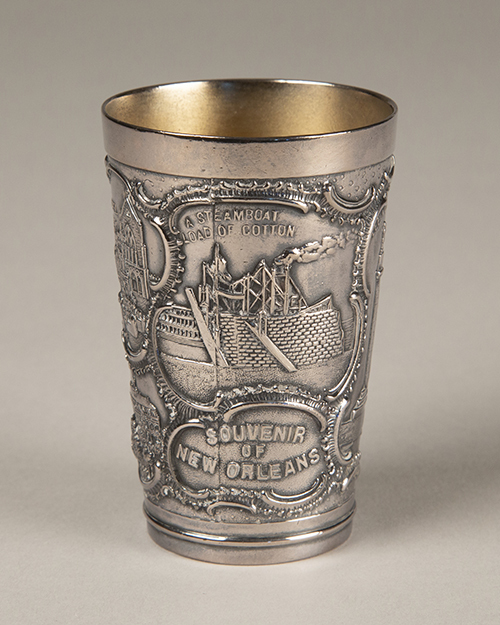 A silver cup intricately engraved with a steamboat and the words A Steamboat Load of Cotton and Souvenir of New Orleans. The cup has ornate detailing around the images and text, giving it an antique appearance.