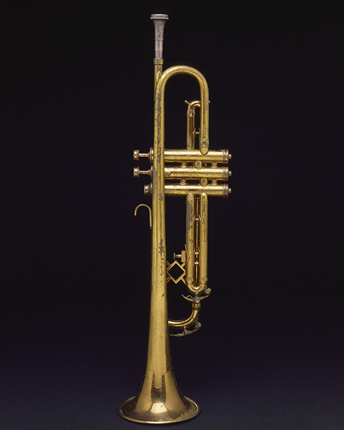 A gold-colored trumpet is positioned upright against a plain black background. Its shiny metallic surface reflects light, highlighting its detailed valves and mouthpiece.