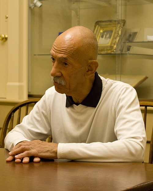 An older person with a mustache sits at a wooden table, wearing a white shirt with a black collar. Bookshelves with various items are in the background.