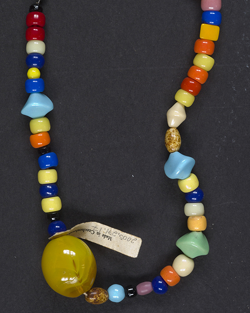 A colorful beaded necklace featuring a large yellow bead. The necklace includes a variety of bead shapes and colors, such as blue, red, green, and orange. A small tag is attached to the necklace. The background is dark gray.