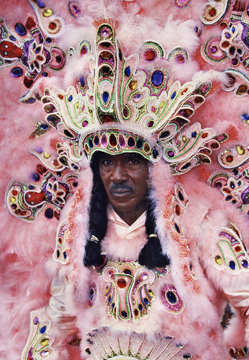 A person wearing an elaborate, colorful costume with intricate beadwork and feathers in shades of pink and peach. The outfit features a large headdress and detailed embroidery, creating a striking and festive appearance.