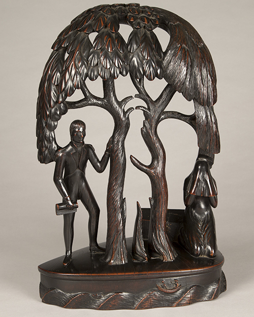 A carved wooden sculpture depicting a man holding a book, standing beside two stylized trees. A seated dog appears near the trees. The artwork has a smooth, dark finish and rests on an oval base with wave-like patterns.
