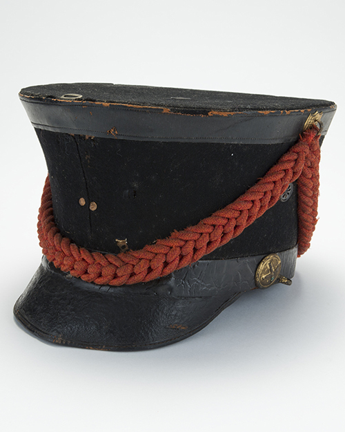 A vintage black military-style cap with a flat top and weathered appearance, featuring a prominent red braided cord across the front and a circular emblem on the side.