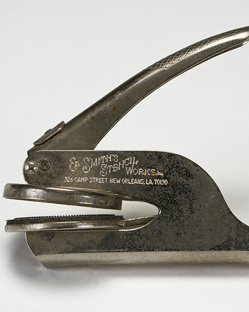 Close-up of a metal embossing seal with the inscription E. Smiths Stencil Works and an address in New Orleans, LA. The tool has a lever for pressing and embossing, designed for creating raised designs on paper.