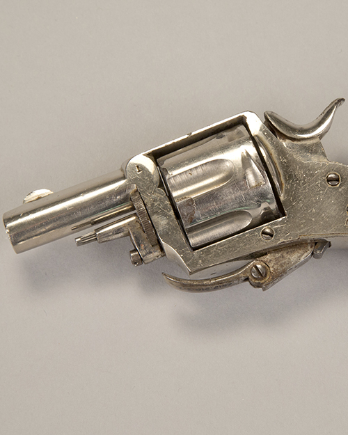 A small, silver, vintage revolver handgun on a plain, light gray background. The revolver is shown from the side, highlighting its cylinder and trigger mechanism.