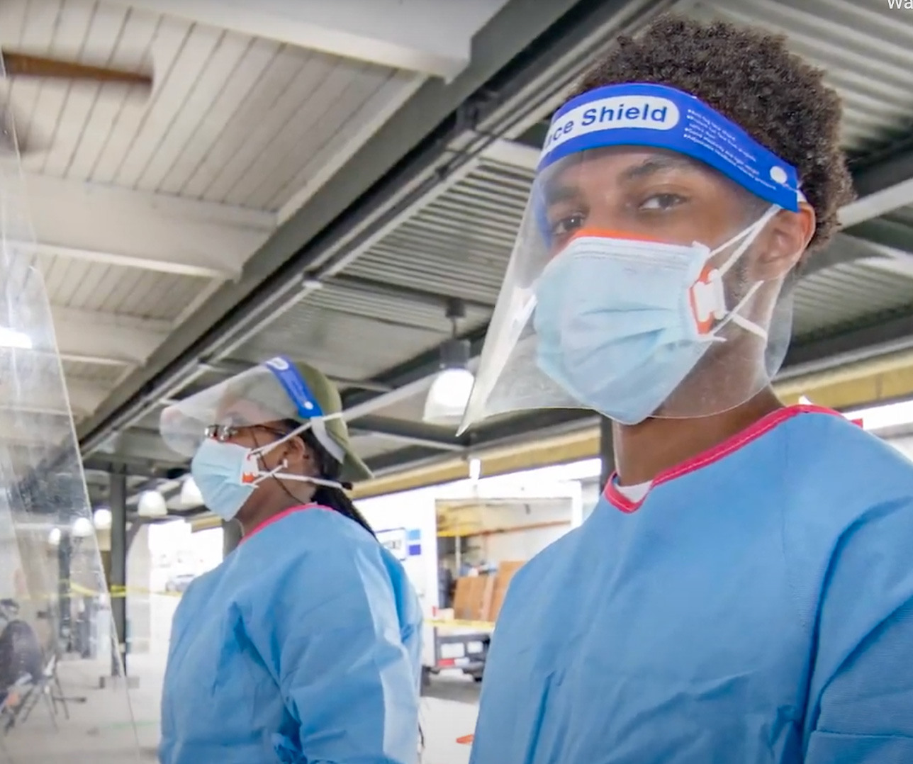 Two healthcare workers wearing blue protective gowns, face shields, and masks stand indoors near a transparent partition. The environment appears to be a medical or testing facility, with visible industrial-style ceilings and fluorescent lighting.