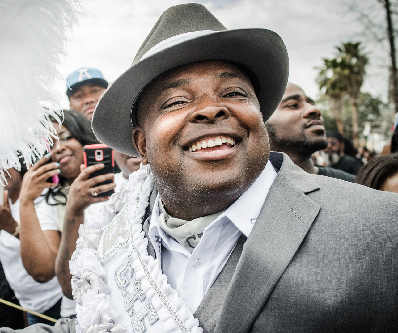 A smiling man in a gray suit and hat participates in a parade. He is surrounded by people, some holding phones to capture the moment. A festive feathered decoration is visible to his left. The background shows a cloudy sky and trees.