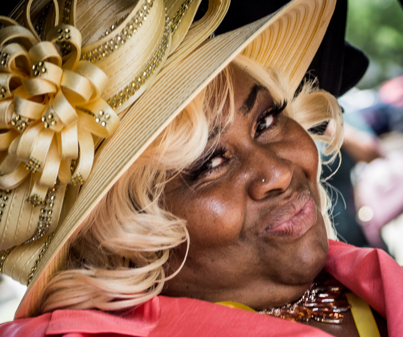 A person smiling warmly, wearing a large decorative hat adorned with ribbons and a flower. They have curled blonde hair and are dressed in a bright, colorful outfit. The background is slightly out of focus.