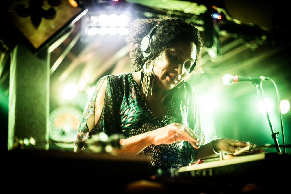A DJ with headphones stands behind a mixing console, immersed in her music. The scene is brightly lit with green lights, creating a vibrant atmosphere. Shes wearing a patterned outfit and large earrings, focused on the equipment.
