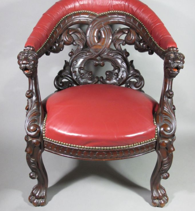 An ornate, antique chair with intricate carved wood featuring lion head details. The seat and curved backrest are upholstered in red leather. The chairs arms and legs display elaborate floral and scroll motifs.