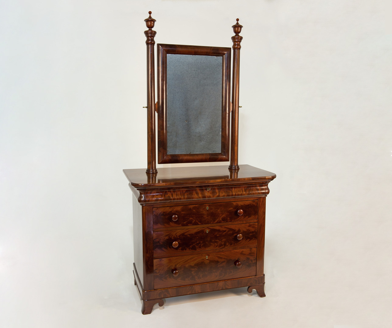 A vintage wooden dresser with four large drawers and ornate handles. It features a tall, rectangular mirror framed in the same wood, supported by two turned posts on either side. The dresser has a polished, dark wood finish.
